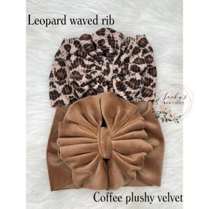 May include: Two baby headbands. The top headband is leopard print with the text "Leopard waved rib". The bottom headband is coffee-colored plush velvet with the text "Coffee plushy velvet". The headbands are on a white, fluffy surface.