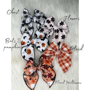 May include: A set of six Halloween-themed hair bows. The bows are made of fabric and feature various designs, including ghosts, bats, pumpkins, flowers, plaid, and a floral pattern with ghosts. The bows are all tied with black ribbon.