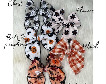May include: A set of six Halloween-themed hair bows. The bows are made of fabric and feature various designs, including ghosts, bats, pumpkins, flowers, plaid, and a floral pattern with ghosts. The bows are all tied with black ribbon.