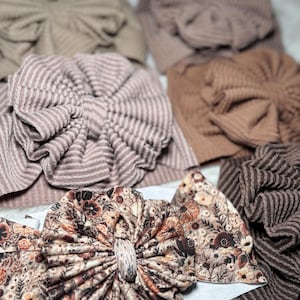 May include: Assortment of baby headbands in various neutral colors. The headbands feature large, decorative bows in solid and striped textures, as well as a floral pattern. The color palette includes shades of brown, tan, and mauve.