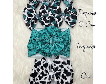 May include: A collection of hair accessories, including several fabric bows and a headband. The bows feature patterns such as cow print, turquoise, and a Carhartt design. The headband is turquoise. The items are arranged on a white, fluffy surface.