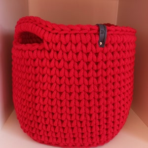 May include: A vibrant red, hand-crocheted basket with a rounded base and two small handles. The basket is made of thick, textured yarn and features a small, dark gray tag with text and a decorative element.