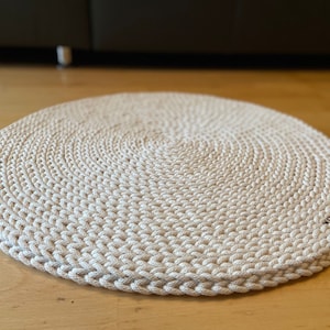 Op de afbeelding: Een rond, crèmekleurig gehaakt vloerkleed met een cirkelvormig patroon. Het vloerkleed is gemaakt van dik, getextureerd garen en heeft een licht verhoogde rand.