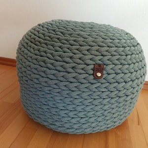 Könnte beinhalten: Ein runder, gewebter Pouf in einem gedämpften Türkis. Der Pouf besteht aus dickem, geflochtenem Stoff und hat ein kleines braunes Lederetikett mit einem Logo. Der Pouf ist ein dekoratives und funktionelles Möbelstück.