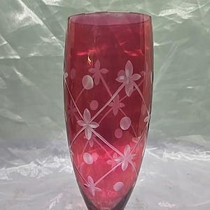 May include: A tall, ruby-red glass with a clear, circular base. The glass features an etched design of flowers and dots, arranged in a diagonal pattern. The glass is approximately 20 cm tall.