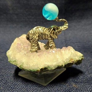 May include: A silver elephant figurine balancing a blue and white marble on its trunk. The elephant is standing on a purple amethyst crystal base.