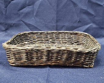 May include: A small, rectangular woven basket with a slightly curved rim. The basket is made of a dark, possibly metallic, material and has a textured appearance. The basket is likely used for storage or display.