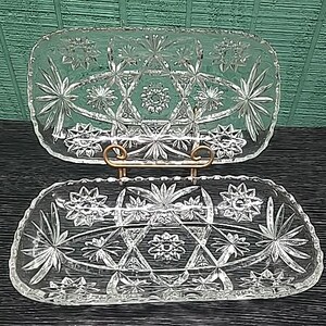 May include: Two clear, rectangular glass serving dishes with starburst and geometric patterns. The dishes are displayed on a gold-coloured metal stand. The background is a dark wood surface with a green textured wall.