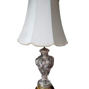 May include: A white ceramic table lamp with a gold base and a white lampshade. The lamp features a detailed floral design with gold accents.