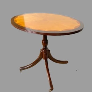 May include: A round, wooden side table with a dark brown trim and a light brown tabletop. The table has a pedestal base with three curved legs and small brass casters. The table is approximately 28 inches in diameter.