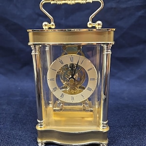 May include: A gold-tone Bulova carriage clock with a rectangular glass case and a top handle. The clock features a circular white face with black Roman numerals, black hands, and a visible internal gear mechanism behind the dial.