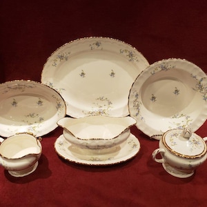 May include: A set of vintage white porcelain dinnerware with a delicate blue floral pattern and gold trim. The set includes a large oval platter, a round bowl, a gravy boat, a creamer, a sugar bowl with lid, and a small oval serving dish.