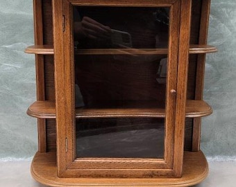 May include: A small, wooden wall-mounted cabinet with a glass-fronted door and two shelves. The cabinet is made of a warm-toned wood and features decorative finials on top. The shelves are rounded and the cabinet has a curved base.