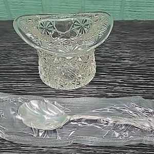May include: A clear glass top hat-shaped container with a detailed, textured pattern. A silver spoon is sealed in a clear plastic wrapper. The spoon has a decorative handle. The items are on a dark surface.