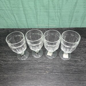 May include: Four clear glass goblets arranged in a row, each featuring a vertical ribbed pattern on the bowl and a sturdy stem. The glasses are viewed from above, showcasing their circular rims and transparent construction on a dark surface.