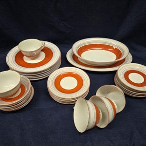 May include: A dinnerware set featuring a white center with a wide burnt orange band and concentric light blue and grey pinstripes. The collection includes stacked dinner plates, salad plates, bowls, and teacups with matching saucers.