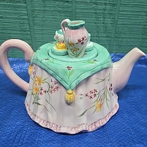 May include: A decorative ceramic teapot with a pink handle and spout. The teapot is painted with floral designs and a green tablecloth design. A small pitcher and other decorative elements sit on top of the lid.