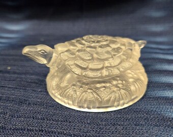May include: A clear glass turtle figurine with detailed shell and body features. The turtle has a detailed head and legs. The figurine is made of clear glass and sits on a blue surface.