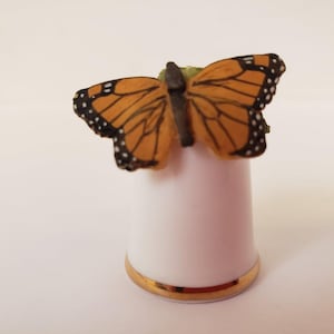 May include: A white ceramic thimble with a gold trim and a detailed butterfly figurine on top. The butterfly has orange wings with black markings.