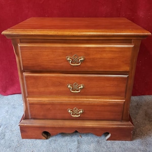 May include: A three-drawer wooden nightstand with a warm cherry finish. The nightstand features ornate brass-colored drawer pulls and a decorative base. The top is rectangular, and the drawers are of equal size. A classic piece of bedroom furniture.
