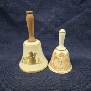 Puede incluir: Dos campanas de porcelana blanca con ribetes dorados y mangos de madera. Una campana presenta un escudo de león dorado, la otra tiene retratos y el texto "CELEBRATE THE WEDDING OF THE PRINCE".