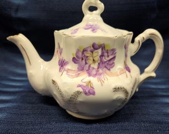May include: A small, white porcelain teapot with a lid, decorated with purple violets and gold trim. The teapot has a curved spout and handle, and a decorative loop on the lid. The base has a textured, dark gray design.