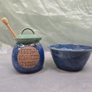 May include: A blue ceramic honey pot with a lid and a wooden dipper, featuring a circular tan medallion embossed with the text APPLE ORCHARD FARM, paired with a matching blue ceramic bowl. Both items have a speckled, glazed finish.