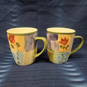 Puede incluir: Dos tazas de cerámica con asas verdes. Las tazas tienen un diseño de patchwork con cuadrados de diferentes colores y flores. Los cuadrados son en tonos de amarillo, azul, verde, morado y rojo. Las flores son rojas y amarillas.