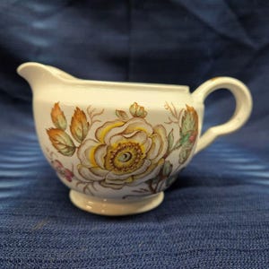 May include: A white ceramic creamer with a floral design. The design features a large yellow flower with brown and green leaves. The creamer has a handle and a rounded base.