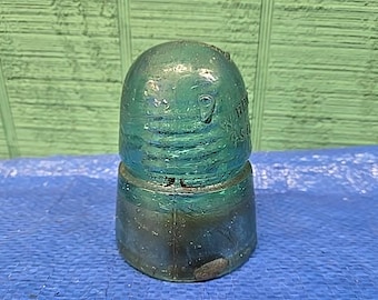 May include: A translucent teal glass electrical insulator with a rounded dome top and a cylindrical base. The glass features embossed text on the side that reads "HEMINGRAY" and "MADE IN U.S.A." with a visible vertical mold seam running down the side.