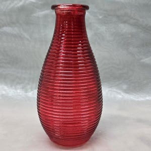Puede incluir: Un jarrón de vidrio rojo con un diseño acanalado. El jarrón es alto y estrecho, con un fondo redondeado y una abertura ligeramente acampanada.