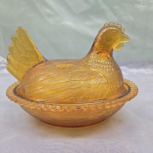 May include: An amber-colored glass hen on a nest dish. The lid is shaped like a sitting hen with detailed feather textures, while the base features a beaded rim and a woven basket-weave pattern. The glass has a translucent, warm orange hue.