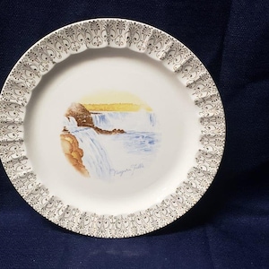 Puede incluir: Un plato de cerámica blanco con un borde dorado y un diseño decorativo. El plato presenta una imagen pintada de las Cataratas del Niágara con el texto "Niagara Falls" debajo de la imagen.