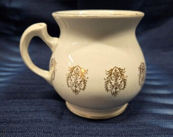 Puede incluir: Taza de cerámica blanca con asa dorada y diseños florales dorados. La taza tiene un cuerpo redondeado y un borde ancho.