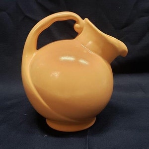 May include: A peach-colored ceramic pitcher with a rounded body and a curved handle. The pitcher has a smooth, glossy finish.