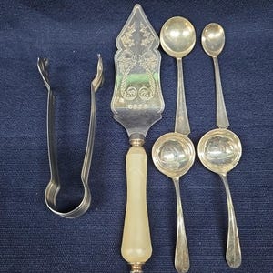 Pode incluir: Um conjunto de servir prateado composto por uma espátula de bolo com cabo em tom de osso e padrões florais gravados, um par de pinças de metal e quatro colheres de tamanhos variados com taças redondas, dispostos sobre um tecido azul escuro.