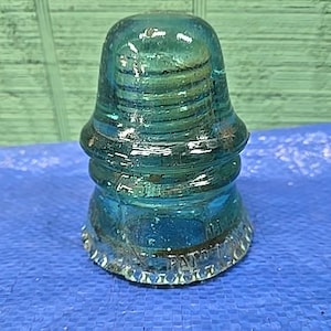 Könnte beinhalten: Ein Vintage-Glasisolator in Teal mit abgerundeter Oberseite und gerilltem Design. Der Sockel hat einen dekorativen, gewellten Rand. Der Isolator besteht aus transparentem Glas und steht auf einer blauen Oberfläche.