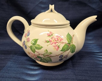 May include: Cream-coloured ceramic teapot with a lid, handle, and spout. The teapot is decorated with pink flowers, green leaves, and purple berries. The teapot sits on a blue surface.