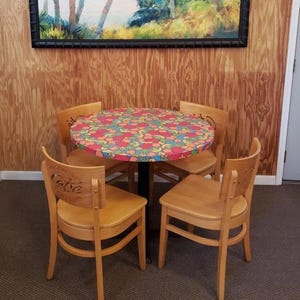May include: A round table with a colorful floral pattern, surrounded by four wooden chairs. The chairs have a curved back and a light brown finish. The table is supported by a black metal base. A painting hangs on the wood-paneled wall.