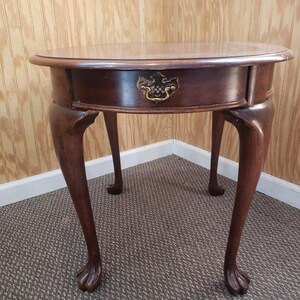 May include: A round, dark brown wooden side table with a single drawer and brass hardware. The table has four curved legs ending in claw feet. The table is approximately 24 inches in diameter and 24 inches tall.