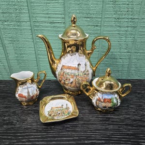 May include: A gold-toned porcelain tea set featuring a coffee pot with a lid, a creamer, a sugar bowl with a lid, and a small square tray. Each piece displays a colorful illustration of Nuremberg cityscapes with the text Nuremberg printed below.