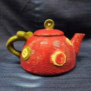 Puede incluir: Una tetera de cerámica roja con asa y tapa verdes. La tetera está decorada con botones coloridos.
