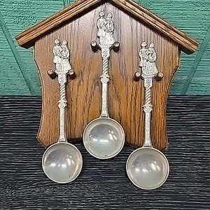 May include: A house-shaped wooden wall plaque featuring three silver-toned spoons with decorative handles. Each spoon handle is topped with a detailed relief figure of a couple in traditional attire. The spoons are mounted on small wooden pegs.