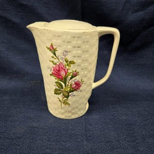 Puede incluir: Jarra de cerámica blanca con textura de cesta tejida y tapa. La jarra presenta un diseño floral con rosas rosas, hojas verdes y detalles morados. Tiene un asa curva y un pico vertedor.