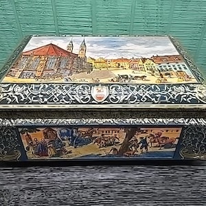 Könnte beinhalten: Eine rechteckige Aufbewahrungsbox aus Metall mit einer farbenfrohen Abbildung eines Marktplatzes mit einer großen Kirche auf dem Deckel. Die Seiten sind mit kunstvollen goldenen und schwarzen Rankenmustern sowie einer Szene mit Menschen verziert.