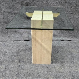 May include: Square glass-top coffee table with a beige stone base and brass accents. The table has a modern design, with a clear glass top and a rectangular base. The brass accents are visible on the top of the base.