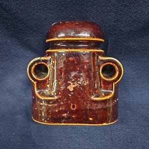 Puede incluir: Un aislante eléctrico de cerámica esmaltada en color marrón oscuro con una parte superior redondeada, un cuerpo central con dos aberturas laterales circulares prominentes y una base plana. El acabado brillante muestra un ligero desgaste.