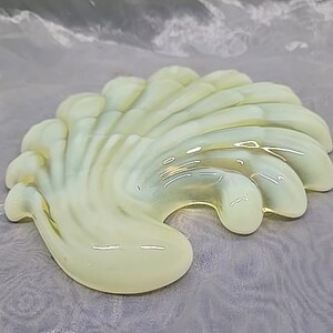May include: A pale yellow, seashell-shaped decorative object. The glossy, translucent piece has a fan-like design with curved ridges and a smooth, rounded base. The object is likely made of glass or a similar material.