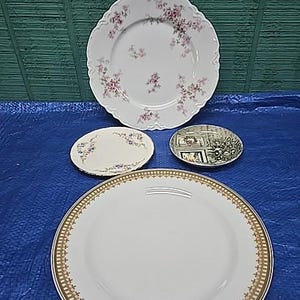 Puede incluir: Cuatro platos decorativos de varios tamaños y diseños. El plato más grande es blanco con un borde dorado. Otro plato tiene un estampado floral, y un plato pequeño presenta un paisaje. Los platos están dispuestos sobre una superficie azul.