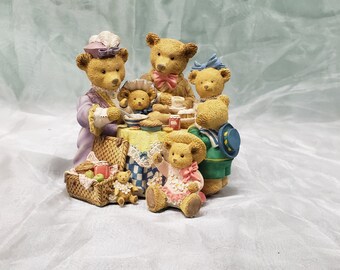Puede incluir: Un grupo de cinco ositos de peluche vestidos con diferentes atuendos se reúnen alrededor de una mesa de picnic. Los osos están hechos de cerámica marrón claro y tienen detalles pintados. La mesa está cubierta con un mantel a cuadros y tiene una cesta de fruta y una jarra de jugo encima. Los osos están todos sonriendo y parecen disfrutar de su picnic.
